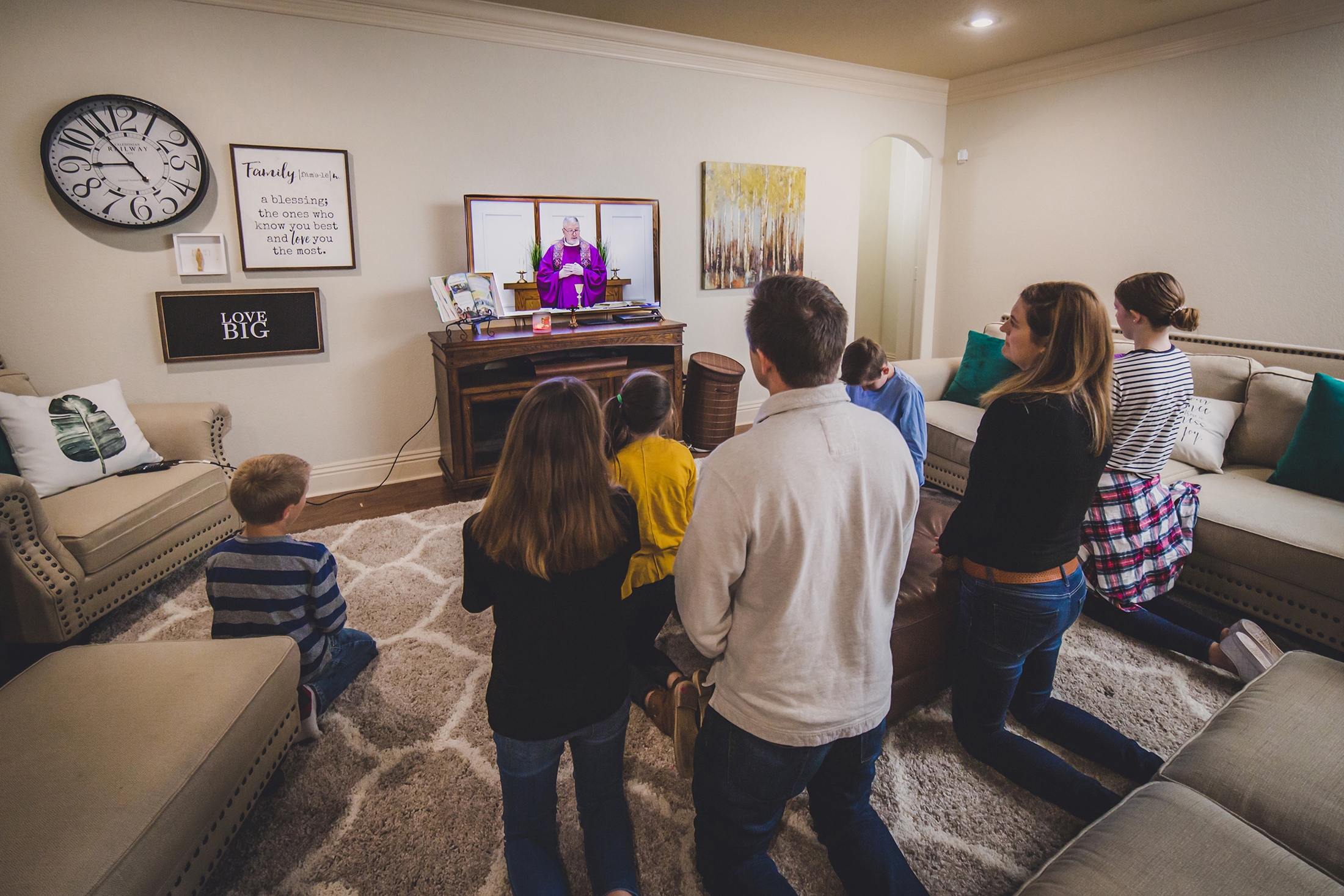 A family kneels in a living room while watching Mass livestreamed on TV