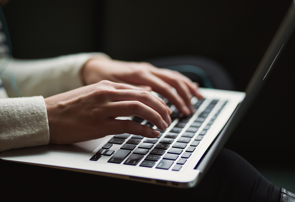 A Caucasian person wearing a white sweater types on a silver-and-black laptop keyboard. Only the hands are visible. 