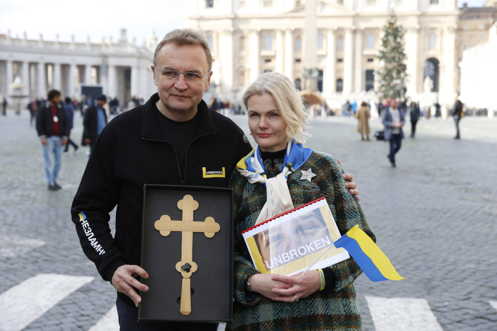 A white man with glasses holds a gold cross next to a white woman with a Ukrainian flag and a picture of a face with the words "unbroken" in St. Peter's Square