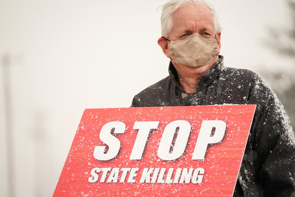 An older, masked man holds a sign that reads "Stop state killing" in the snow