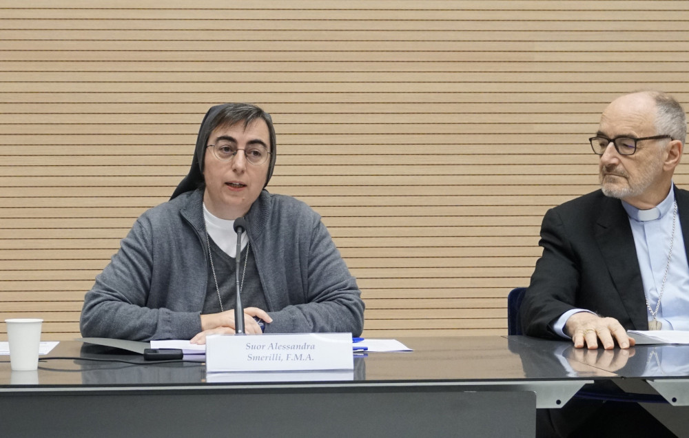 A white sister wearing a grey habit sits next to a white cleric wearing a suit