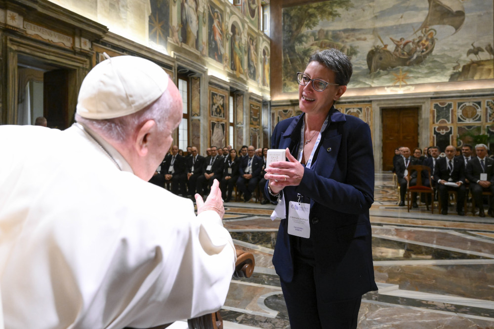 A white woman with short hair and glasses stands in front of Pope Francis with a large group behind her