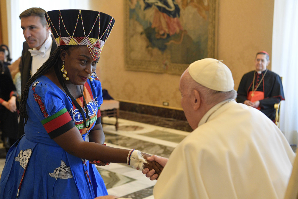 A Black woman wearing a colorful dress and a headdress shakes Pope Francis' hand