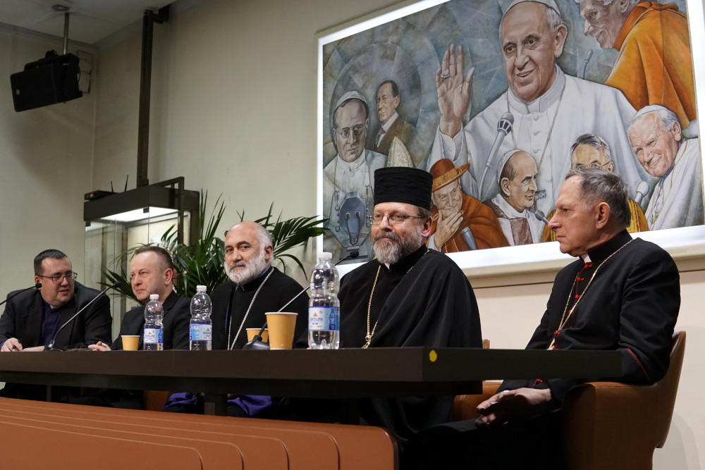 Five white men in black religious garb sit behind a long table with microphones