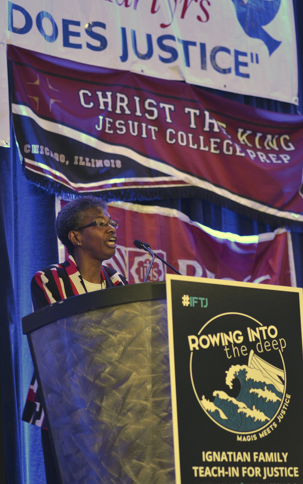 An older Black woman with glasses stands behind a podium with an "Ignatian Family Teach-In for Justice" poster on the front