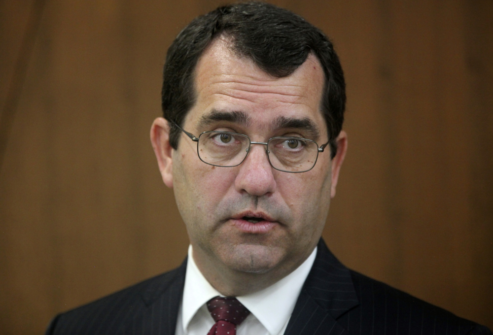 A headshot of pale-skinned man with brown hair and glasses wearing a suit and tie