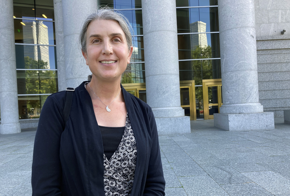 An older white woman wearing a cardigan stands outside a building with white marble columns