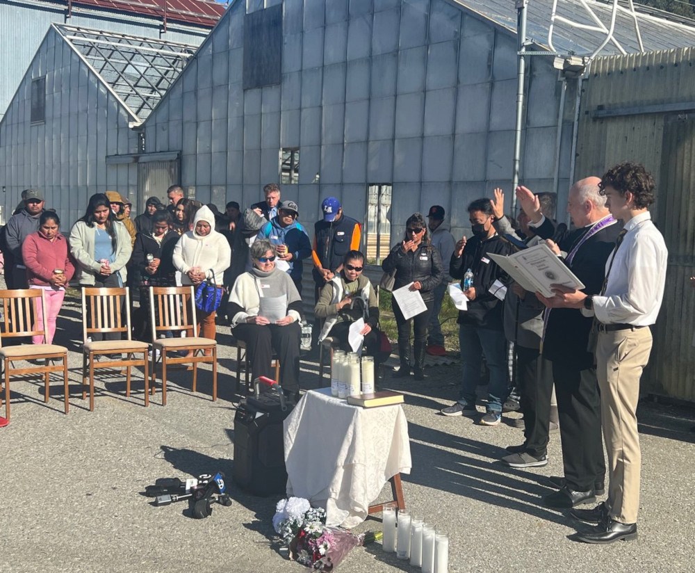 A group of people stand outside greenhouses around a table with a white cloth, many candles, and flowers