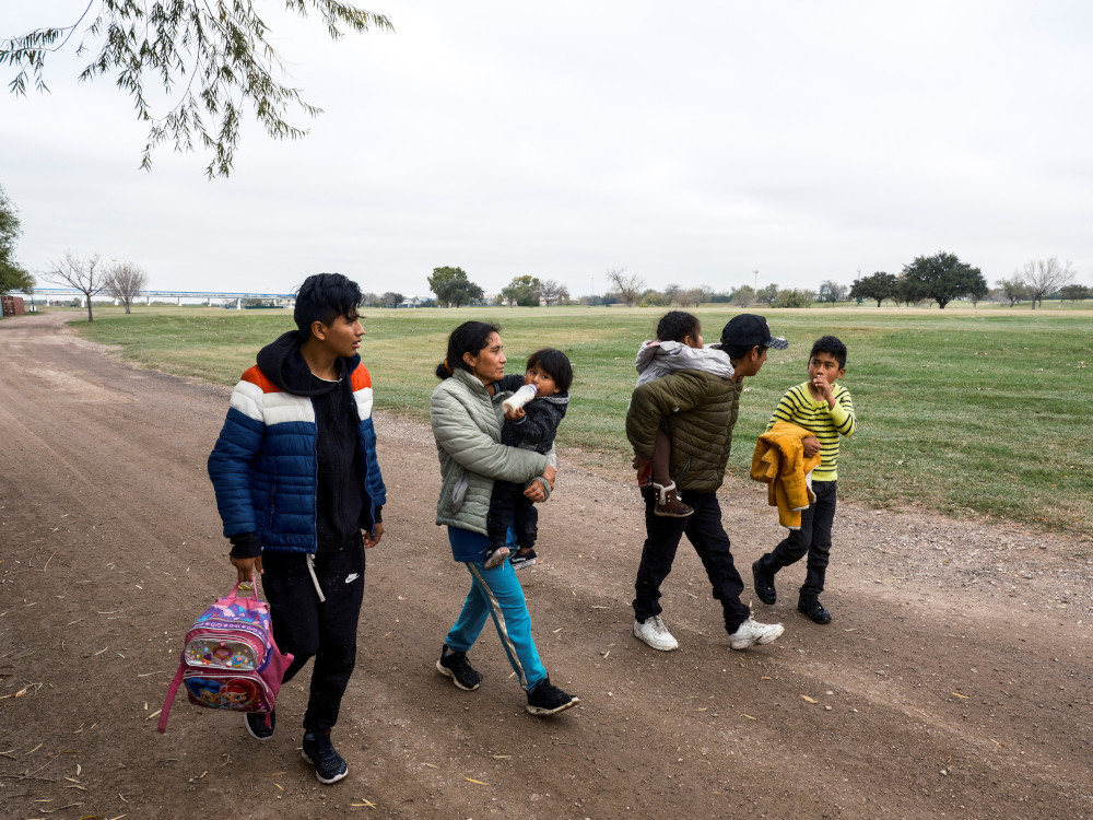 A family with adults carrying children walks along a dirt road next to a grass fiedl