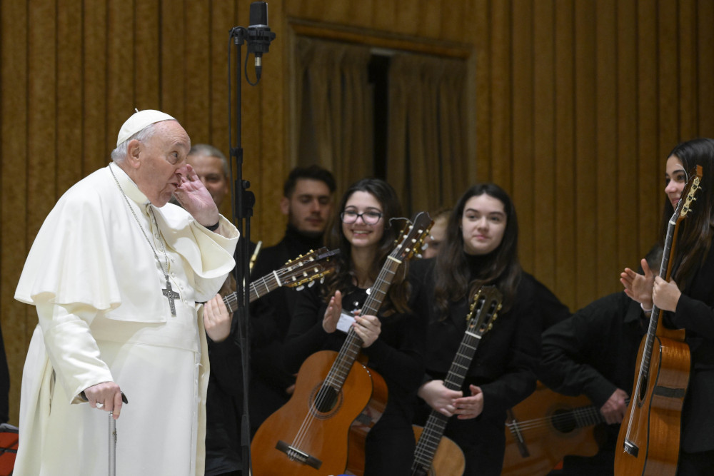 Pope Francis uses a cane and puts his hand to his ear as he stands in front of a group of young guitar players