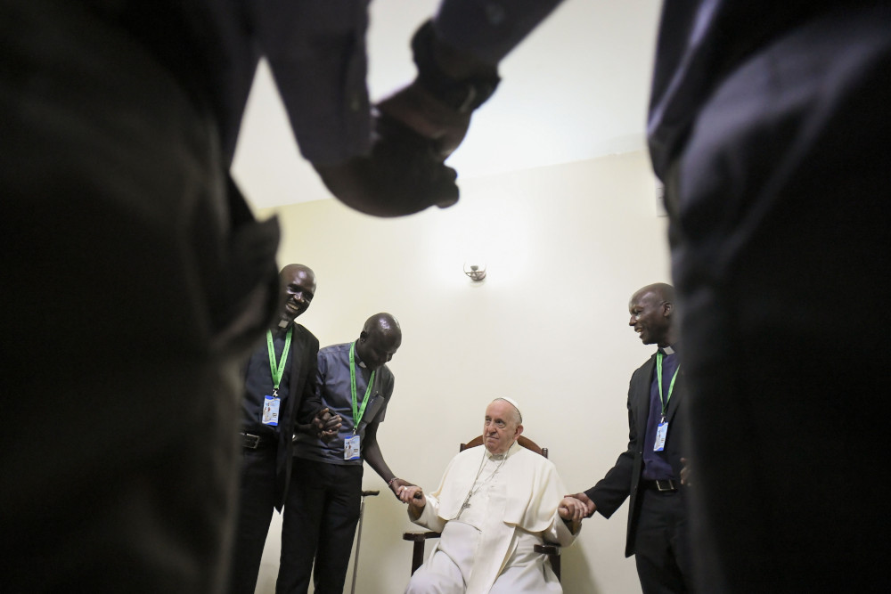 Pope Francis, sitting, holds hands with African Jesuits standing in a circle