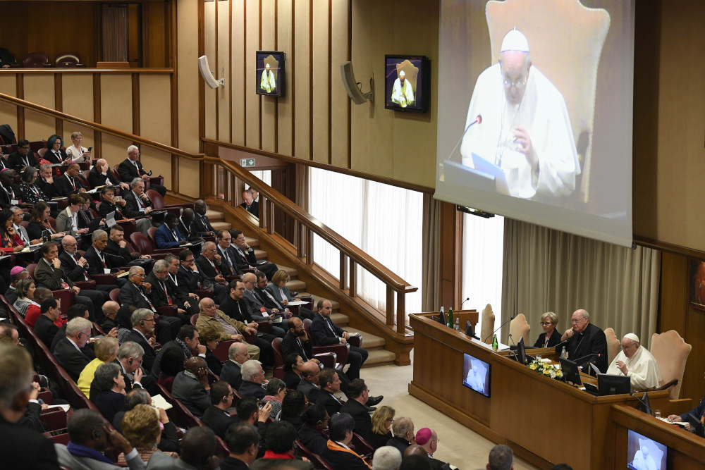 Pope Francis is projected on a large screen in front of a hall of people