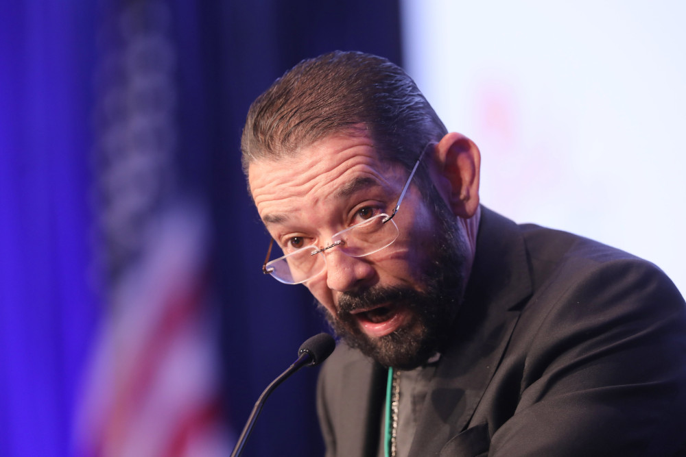 A black-haired man with glasses and a beard wears a black cassock and jacket and speaks into a microphone