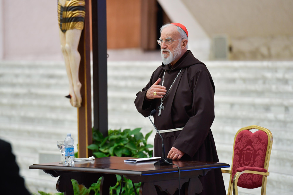 A older white man with a beard and glasses wears a brown tunic and red zucchetto and stands by a crucifix