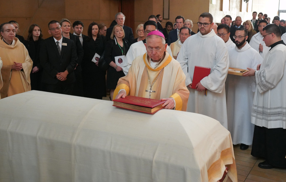 Archbishop Jose Gomez holds a red book over a casket draped in white. Many lay people and priests stand behind him.