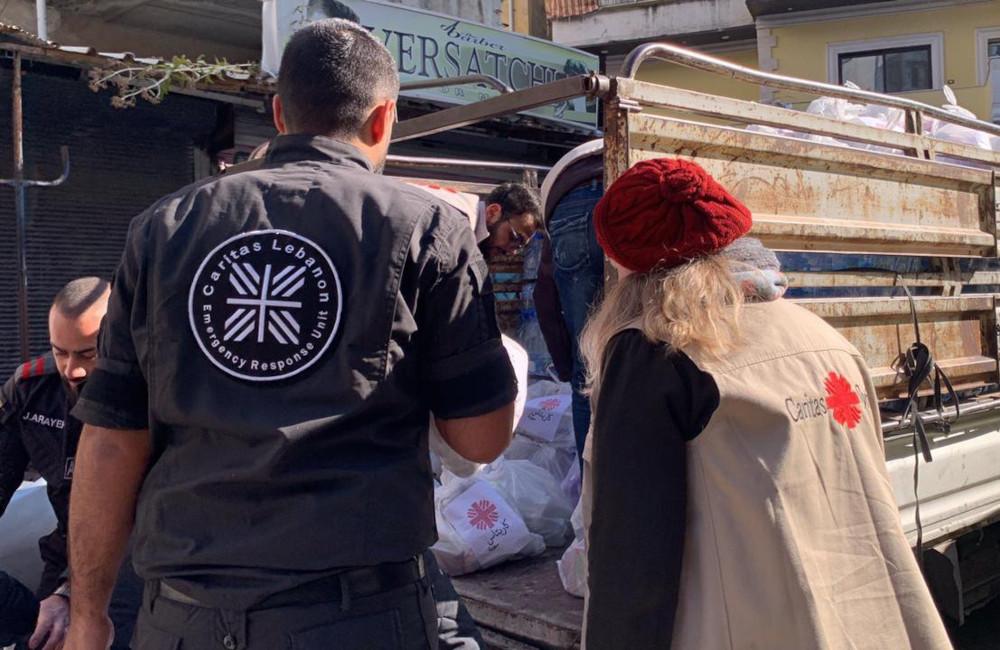 Two people whose backs face the camera wear Caritas jackets. They face a truck.