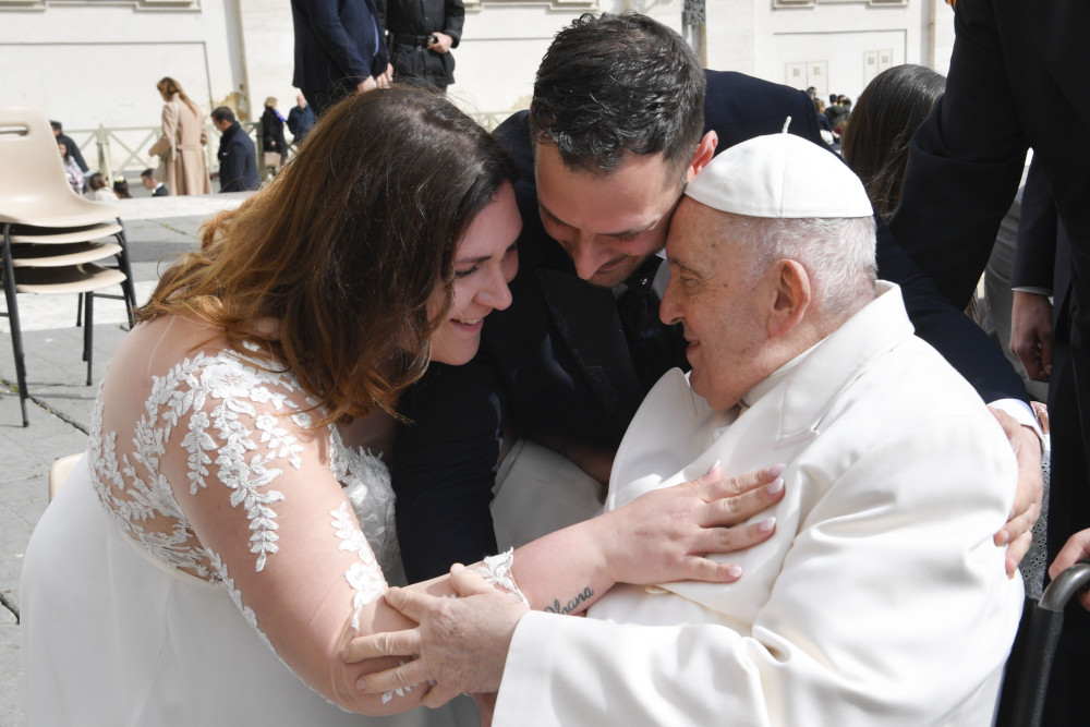 A woman in a wedding dress and a man put their heads close to Pope Francis, who is sitting in a wheelchair