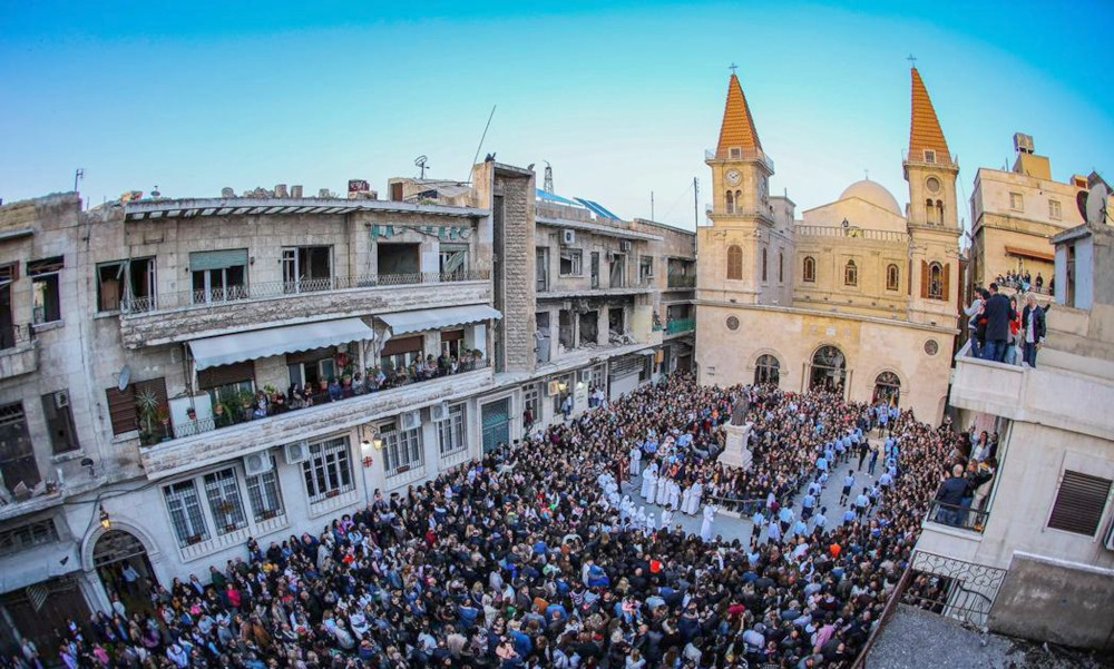 A crowd gathers in a plaza in front of a steepled church. Other people watch from apartment balconies.