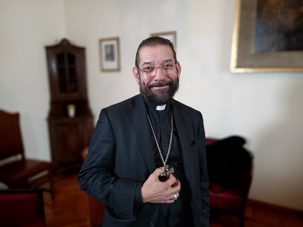 A bearded black-haired man wearing a black cassock, a black jacket, and pectoral cross smiles at the camera