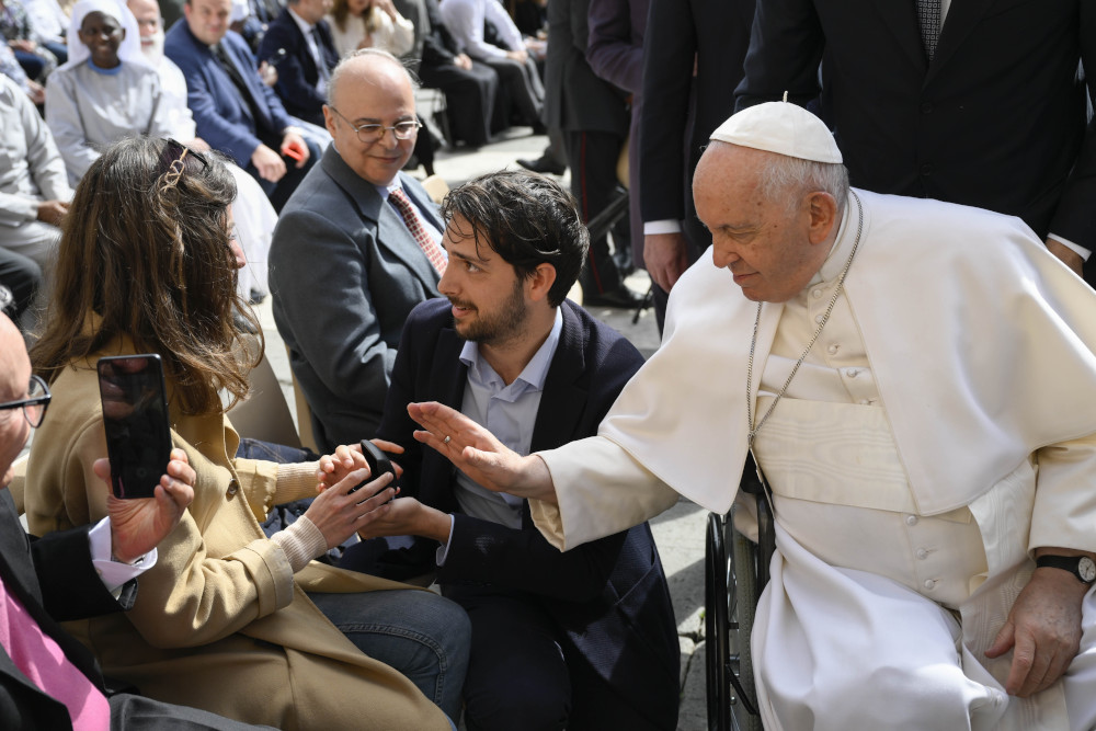 Pope Francis, seated in a wheelchair leans his hand toward a couple, where a man is kneeling with a ring box