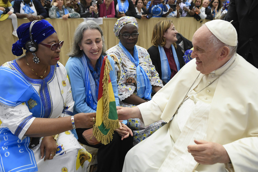 Pope Francis sits in front of a row of women dressed in blue and reaches out to touch a red, yellow and green flag one woman carries