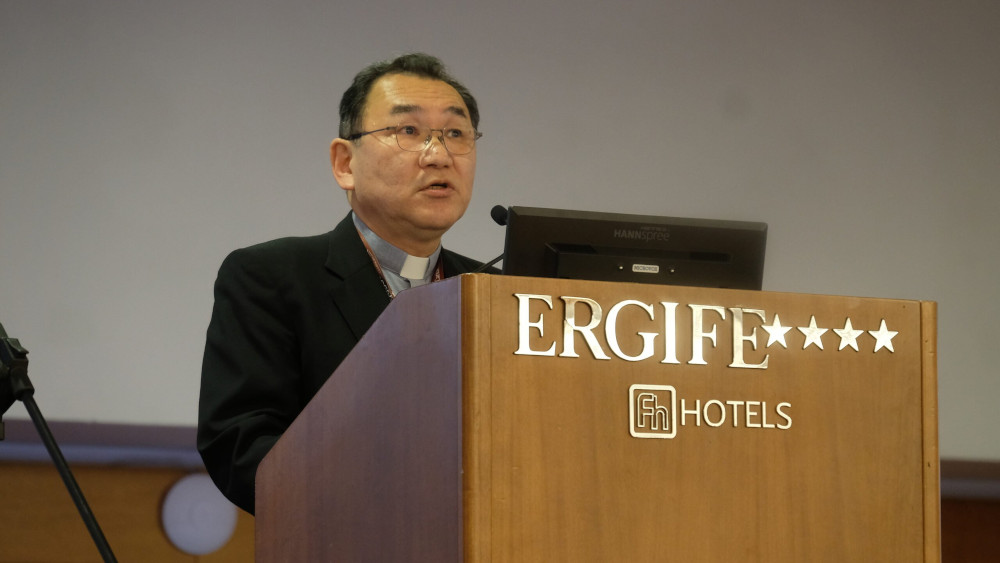 An East Asian man with a clerical collar speaks behind a podium labelled "Ergife Hotels"