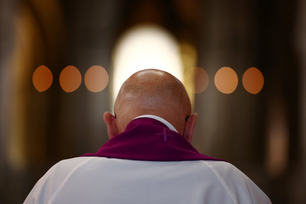 A bald person is seen from behind wearing white and a stole and bowing their head 