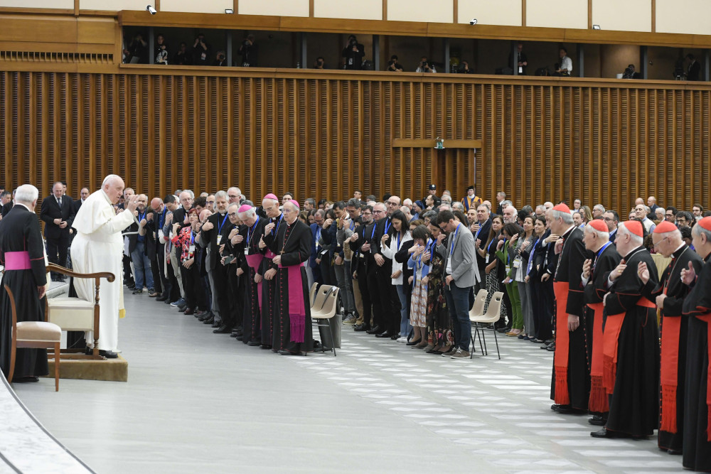 Pope Francis stands and raises his hand in front of a crowd that includes people dressed in the garb of cardinals, bishops and laypeople