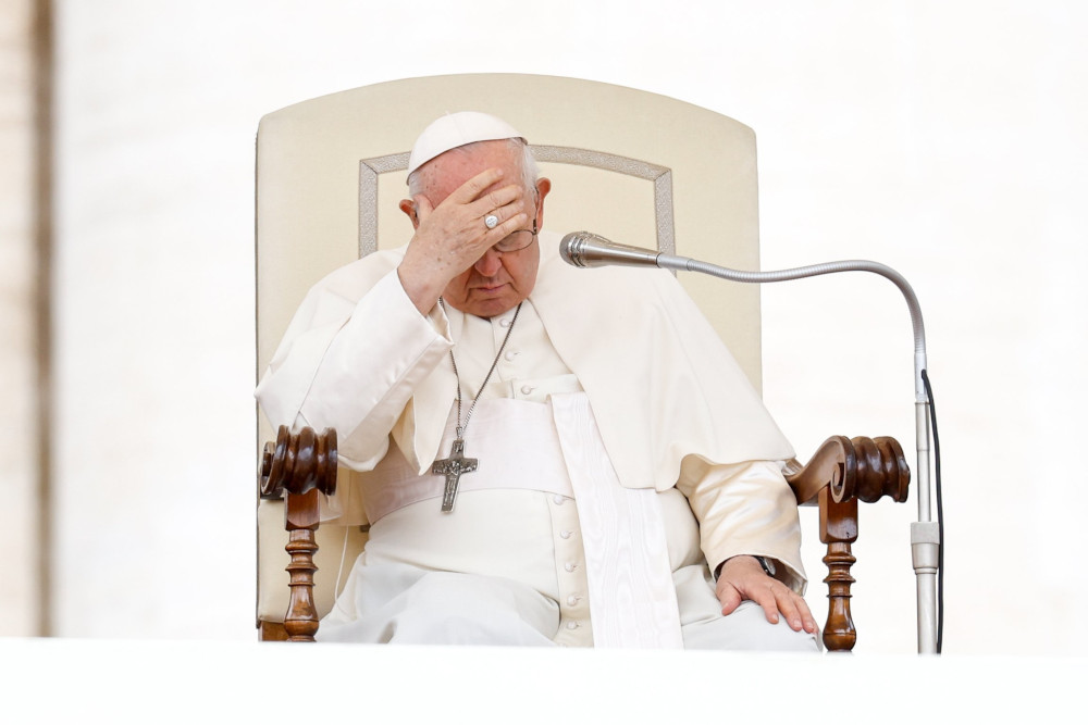 Pope Francis looks down and places his head on his forehead as he sits behind a microphone