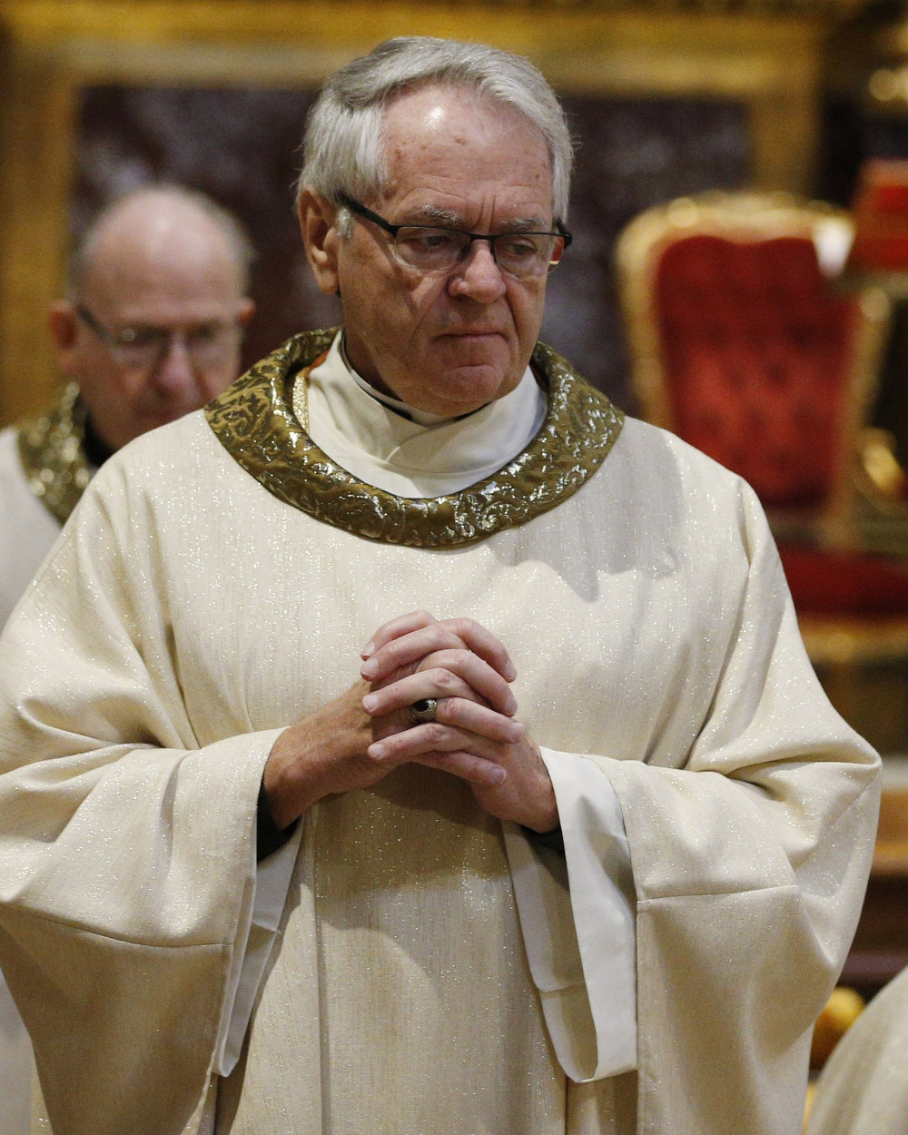 A white man with glasses wears a white and gold chasuble and folds his hands in front of him