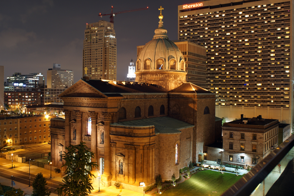 A cathedral with a dome and columns is lit up at might with tall buildings surrounding it