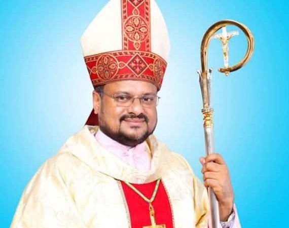 A light-skinned Indian man wears a red and white mitre and holds a crozier