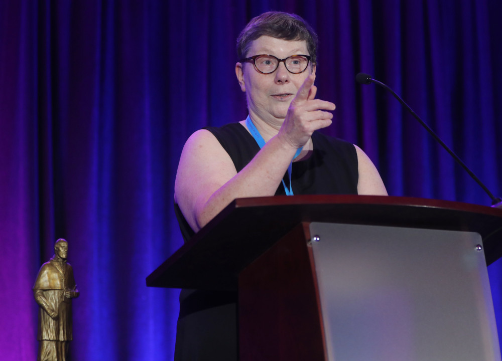 A white woman with short hair and glasses stands behind a podium and points as she is speaking