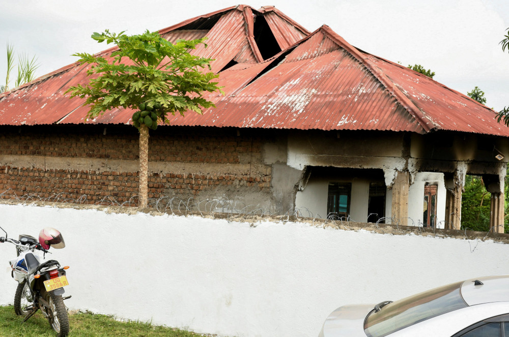 A building with an orange roof and brown brick sides shows black marks and gaps in the ceiling. It stands behind a white wall with barbed wire.