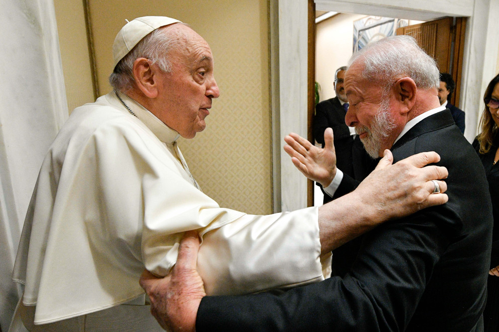 Pope Francis grips the shoulder of an older, white-appearing man in a suit, who in turn grips Pope Francis' upper arm