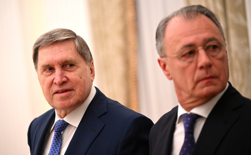 A white man wearing a suit and blue tie stands next to another white man wearing a suit