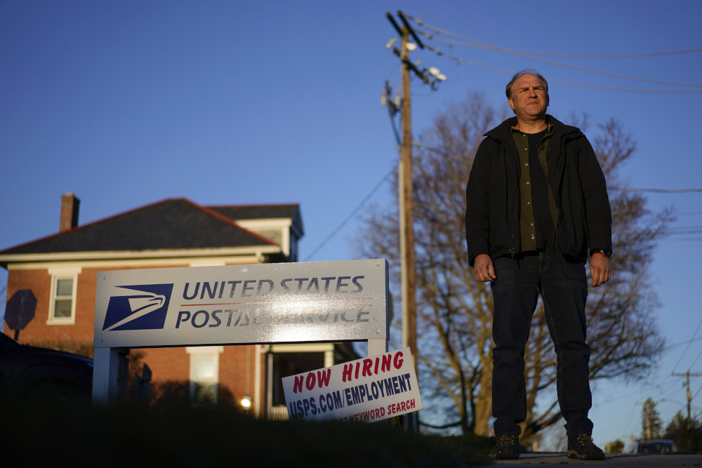 A white man wearing street clothes stands outside a United States Postal Office