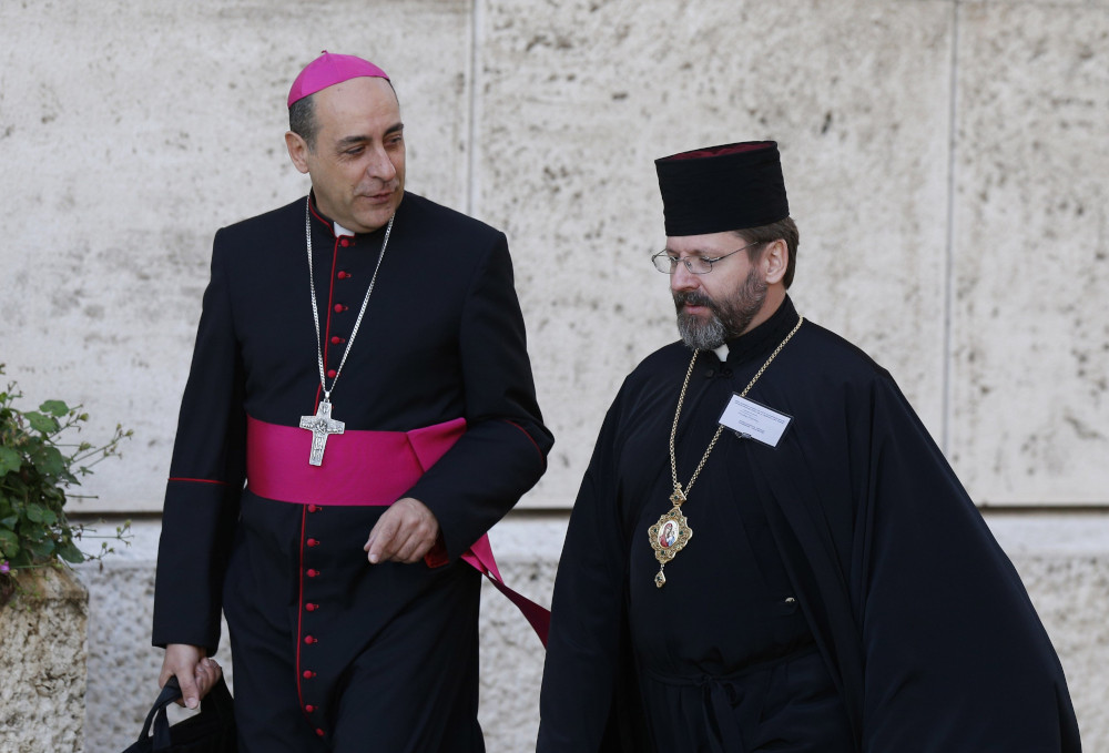 A man wearing a violet zucchetto stands next to another man wearing a skufia hat