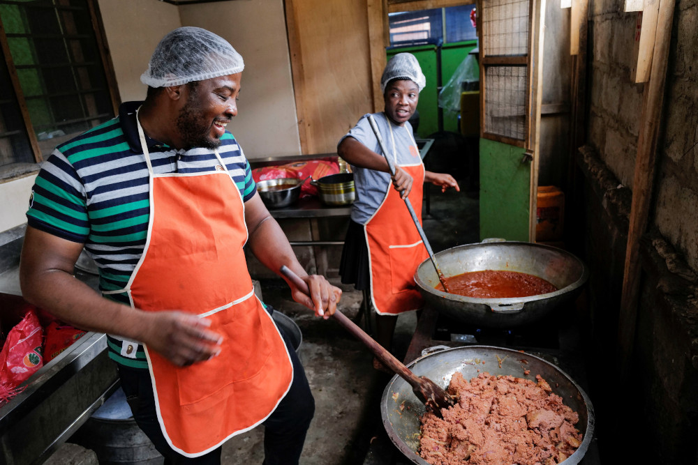 Two Black people wearing hair nets and orange aprons smile and stir food in large pots