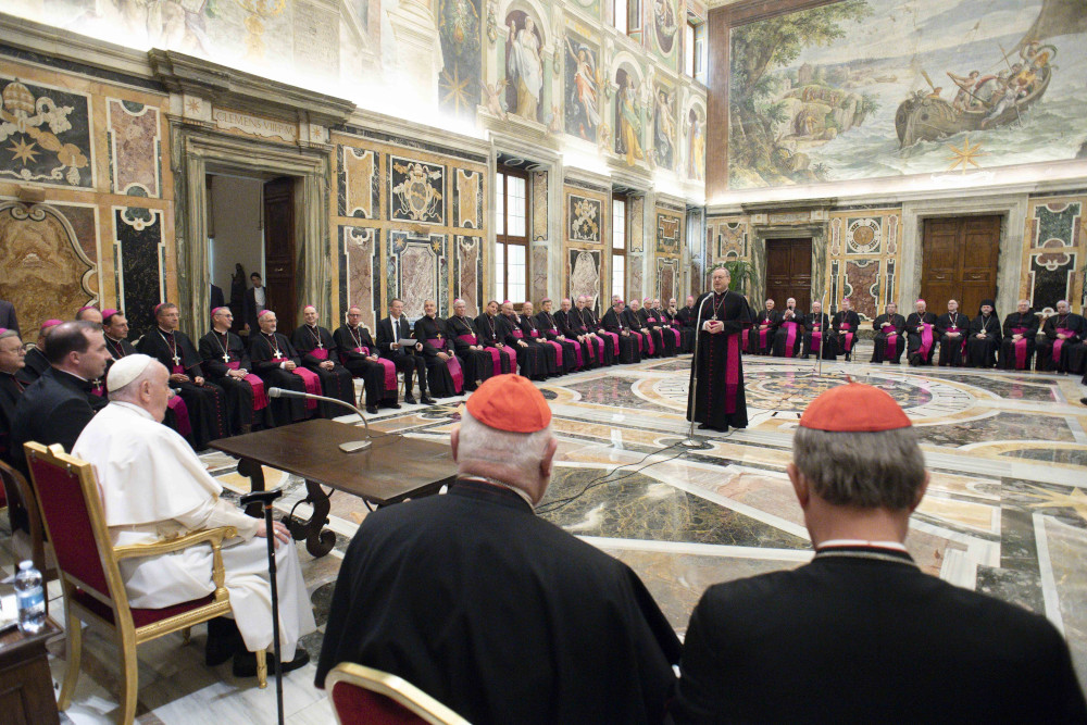 A man wearing a violet zucchetto stands in the middle of a room covered in frescos as he addresses Pope Francis and other bishops and cardinals