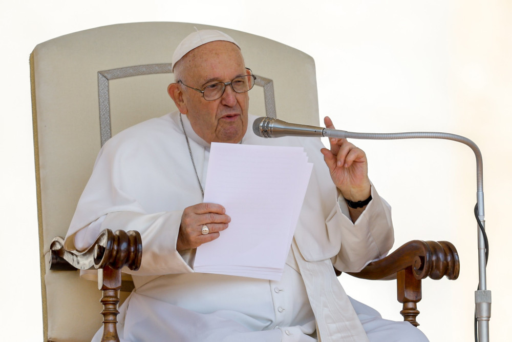 Pope Francis sits in a tall-backed white chair, holds a paper, speaks into a microphone and makes a pointing gesture