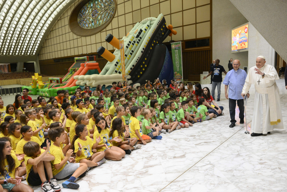 Pope Francis waves and uses a cane to walk across the front of a large group of children wearing t-shirts seated on the floor. An inflatable Titanic and other inflatable slides are visible in the background