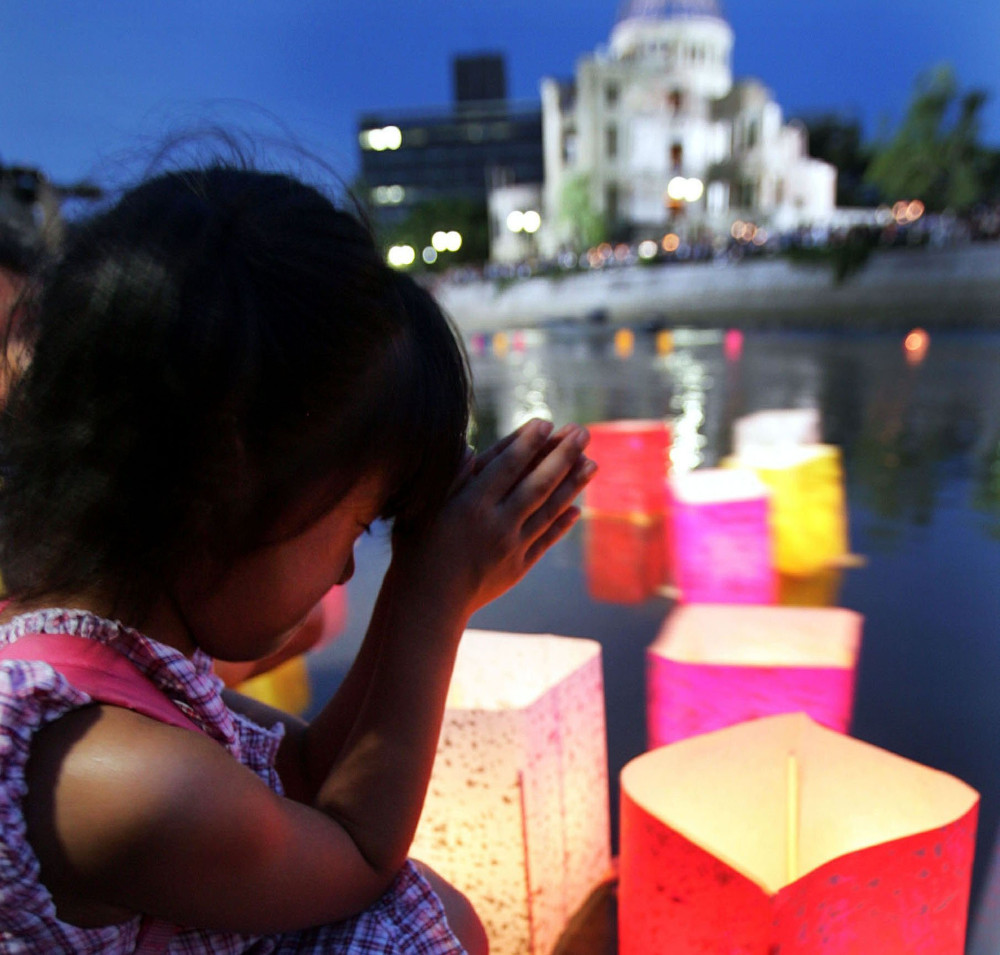 A Japanese child squats and presses her palms together and thumbs to her forehead in front of a waterway with floating paper lanterns. A white building is blurry in the background