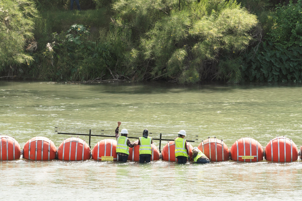 Three men in bright yellow vests address large orange buoys spaced closely in the river