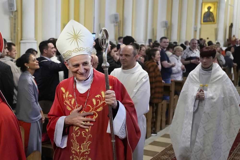 A man wearing a white and gold mitre and red vestments walks down a church aisle