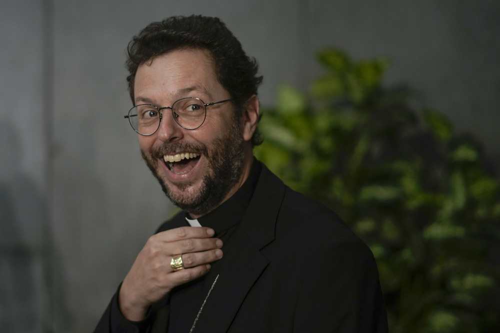 A younger man wearing glasses and a clerical collar beams at the camera while touching his collar