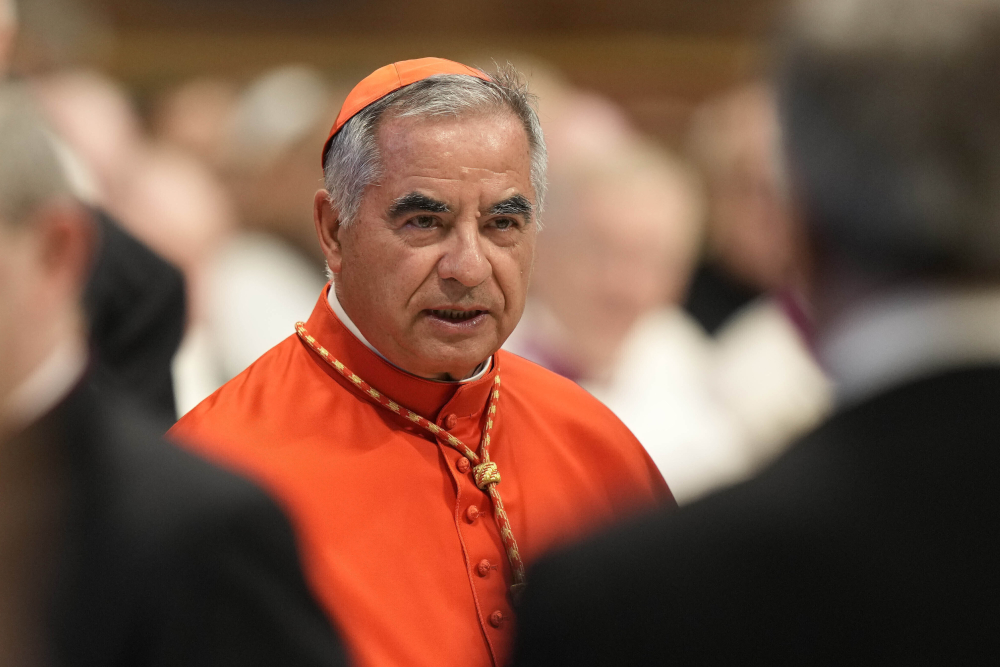 An older man wearing a red zucchetto and a red choir dress vestment stands amid blurred out people