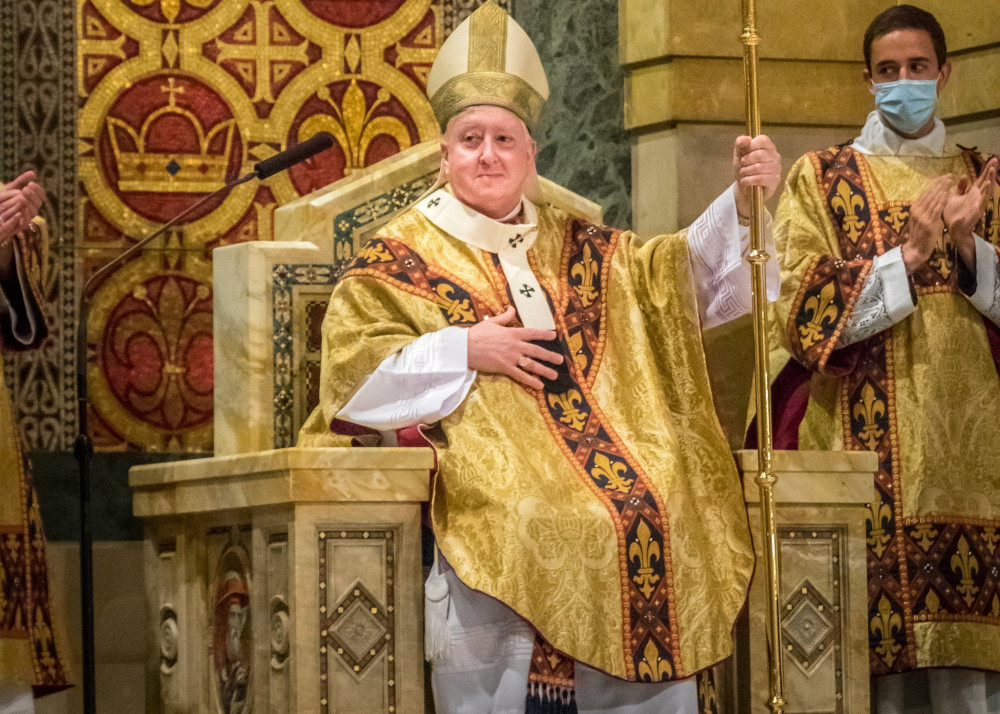 A white man wears a gold mitre and vestments and sits in an ornate chair holding a crozier