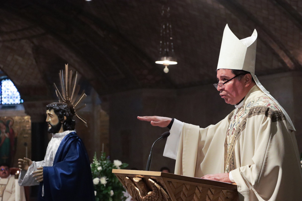 A man wearing glasses and white mitre and vestments stands at a lectern and speaks with an outstretched hand