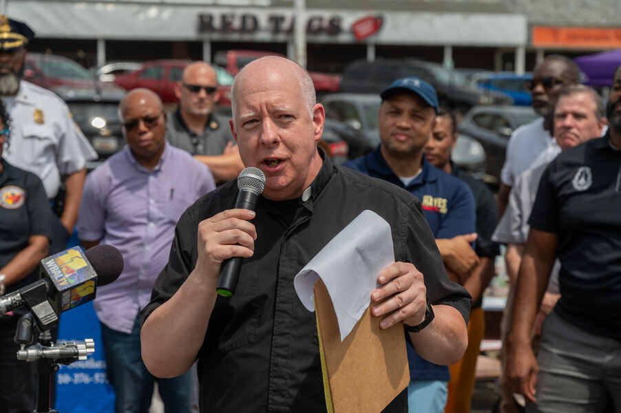 A white man wearing a clerical collared shirt speaks into a microphone as men stand behind him